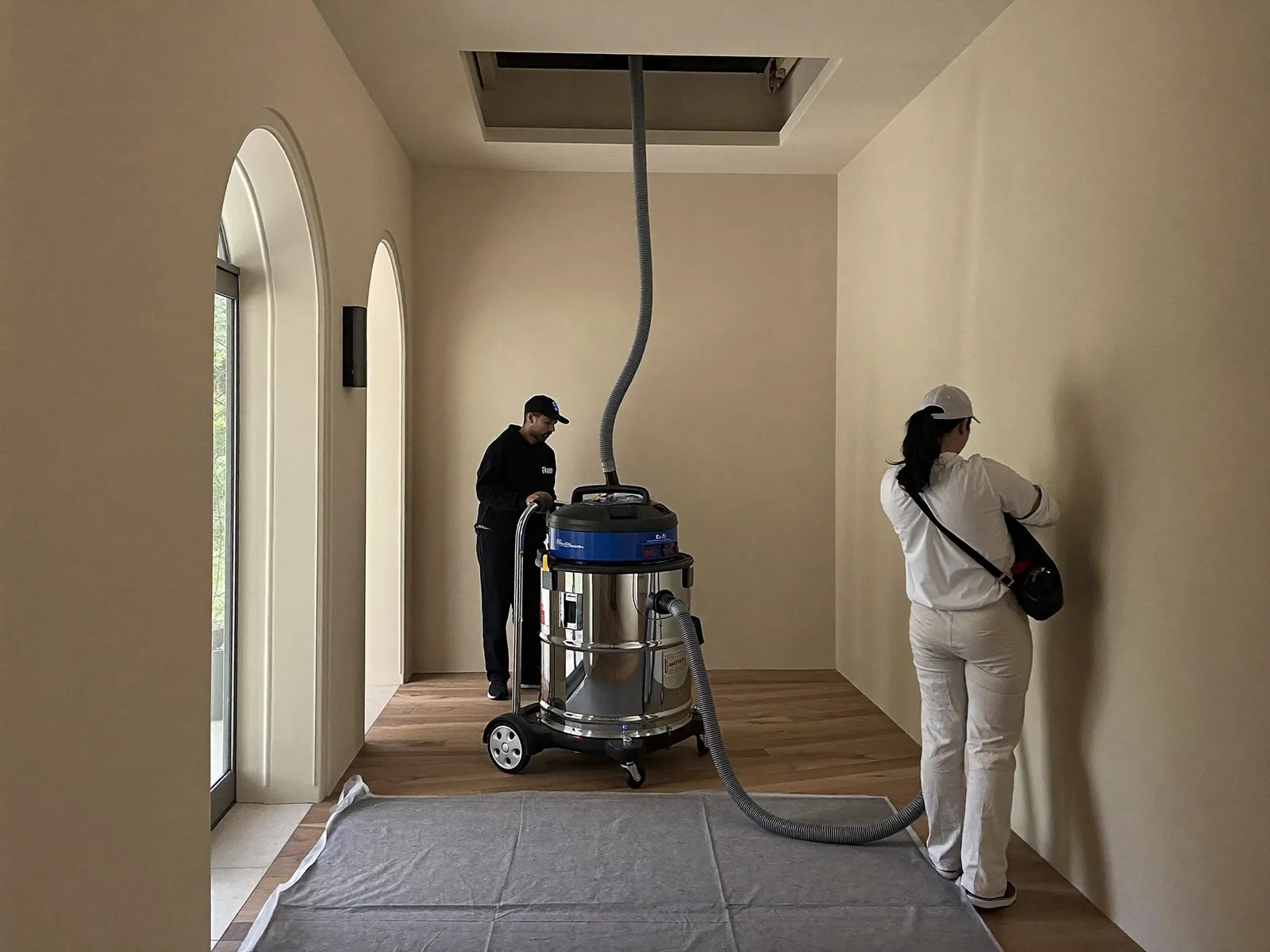 Two technicians connecting a duct vacuum hose to an open ceiling access panel in a villa corridor